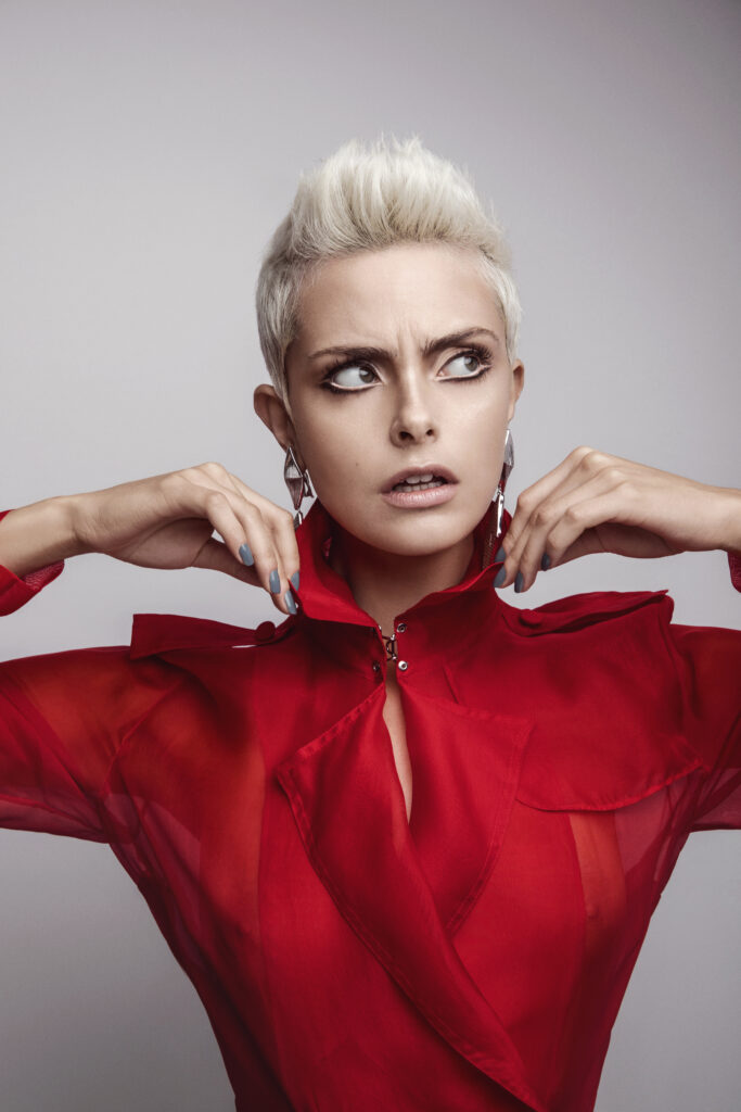 retouching portfolio example of close-up portrait of a woman with short platinum-blonde hair wearing a sheer red high-collared blouse and large silver earrings.