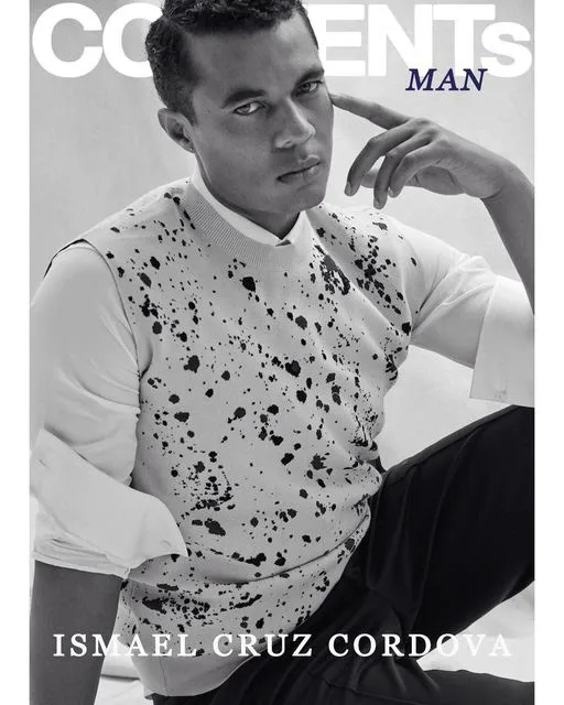 Black and white editorial portrait of Ismael Cruz Cordova for CONTENTS MAN magazine, wearing a white paint-splattered sweater vest over a white shirt, posing with hand near face in a confident studio setting.