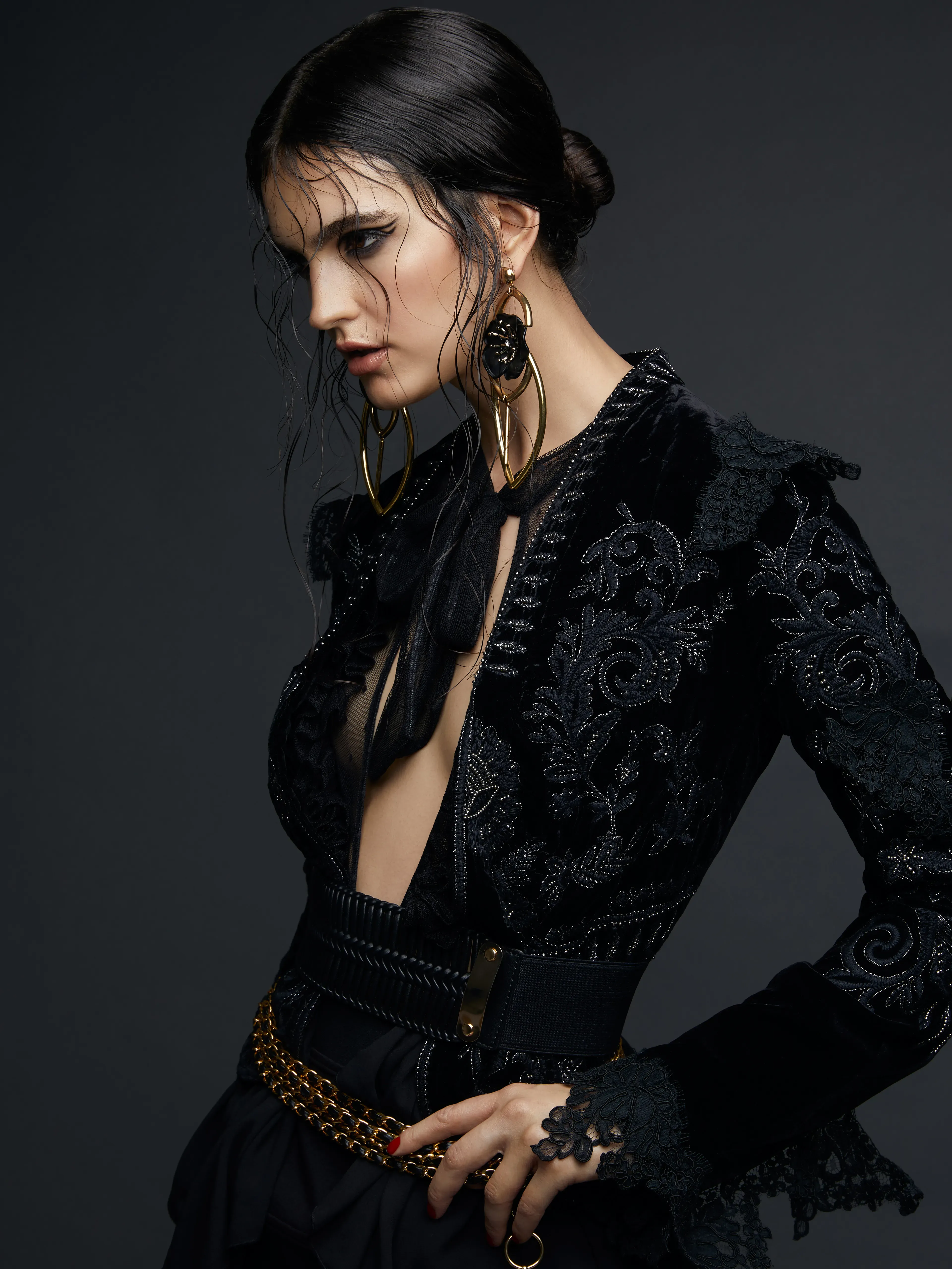 Dramatic fashion portrait of a model in an ornate black velvet jacket with intricate lace and embroidery details, bold gold earrings, and wet-look hair pulled back.