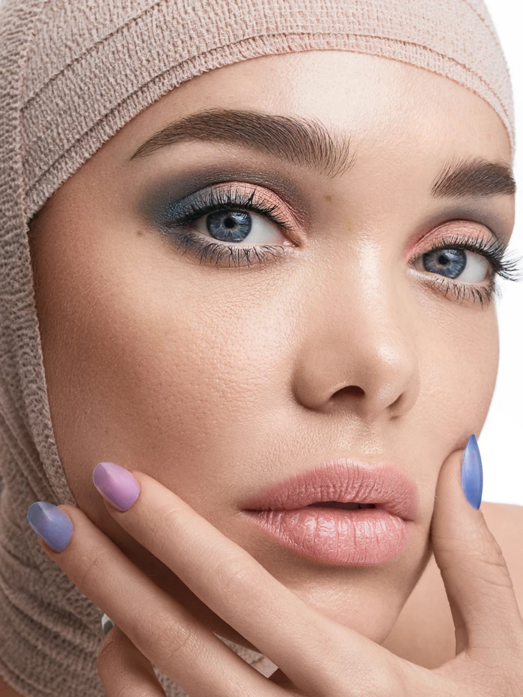 Close-up portrait of a woman with blue eye makeup and pink lips, her head wrapped in beige medical bandage.