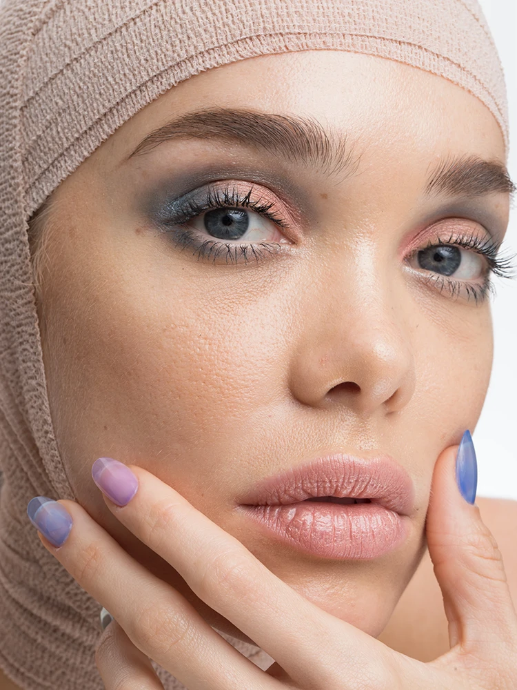 Close-up portrait of a woman with blue eye makeup and pink lips, her head wrapped in beige medical bandage.