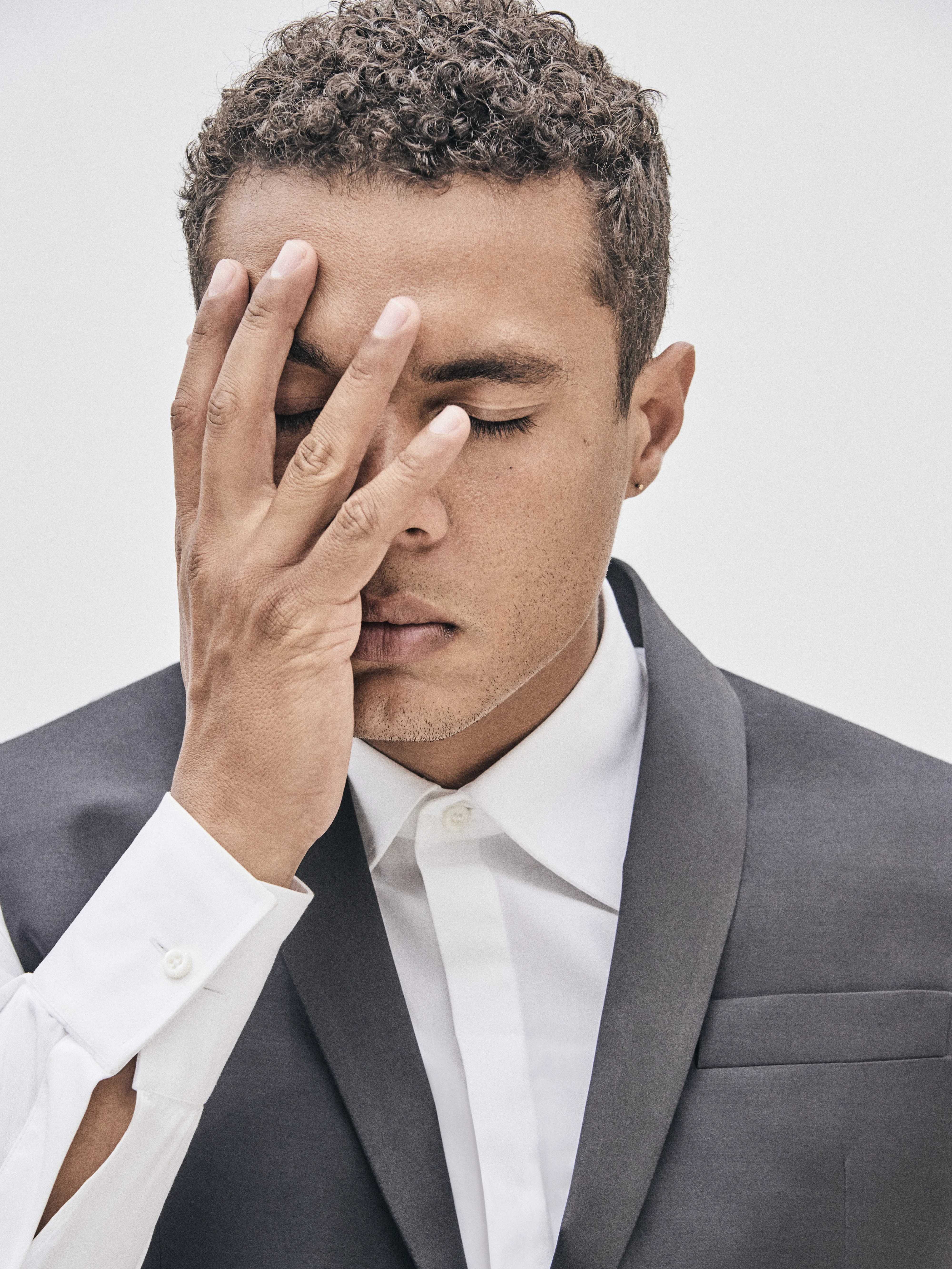 Close-up studio portrait of actor Ismael Cruz Córdova with his hand covering his eyes, wearing a gray suit and white shirt, soft lighting, contemplative expression.