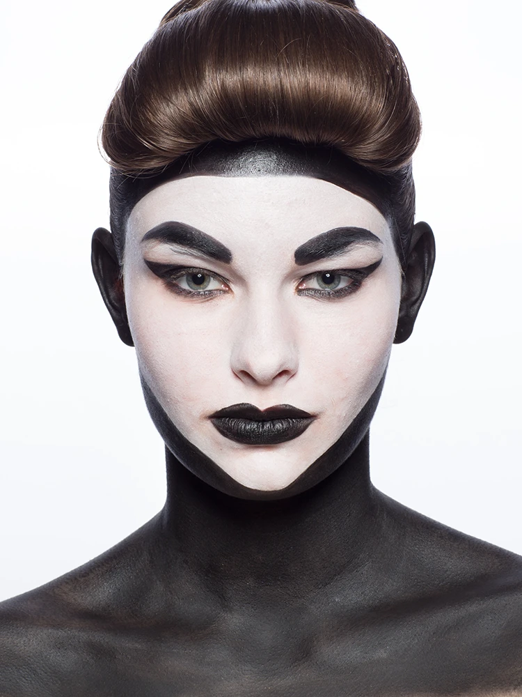 Close-up portrait with high-contrast black and white face paint, geometric brows, and sculpted brown hair.