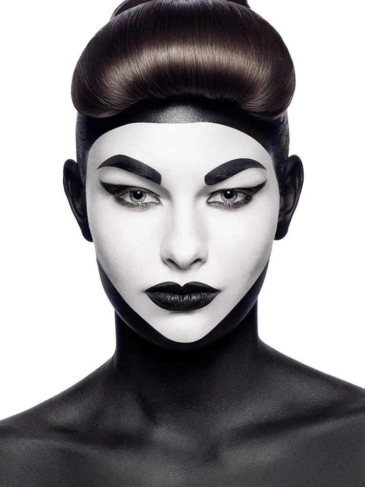 Close-up portrait with high-contrast black and white face paint, geometric brows, and sculpted brown hair.